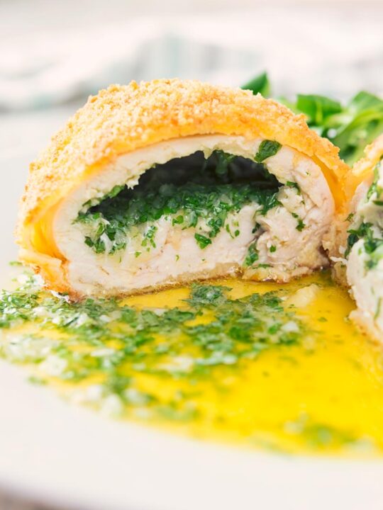Close up portrait image of a garlic chicken Kiev cut open releasing it's garlic butter and parsley all over a white plate, served with a side salad and mashed potato