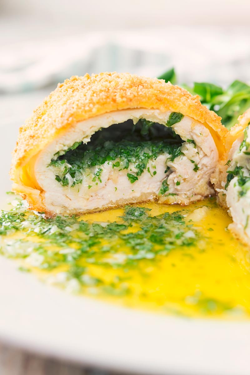 Close up portrait image of a garlic chicken Kiev cut open releasing it's garlic butter and parsley all over a white plate, served with a side salad and mashed potato
