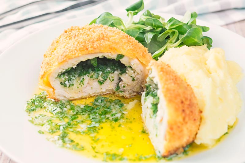Landscape image of a garlic chicken Kiev cut open releasing it's garlic butter and parsley all over a white plate, served with a side salad and mashed potato