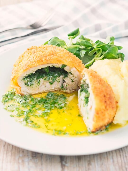 Portrait image of a garlic chicken Kiev cut open releasing it's garlic butter and parsley all over a white plate, served with a side salad and mashed potato