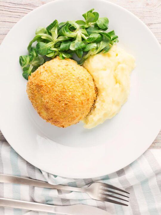 Portrait overhead image of a crispy garlic chicken Kiev on a white plate, served with a side salad and mashed potato
