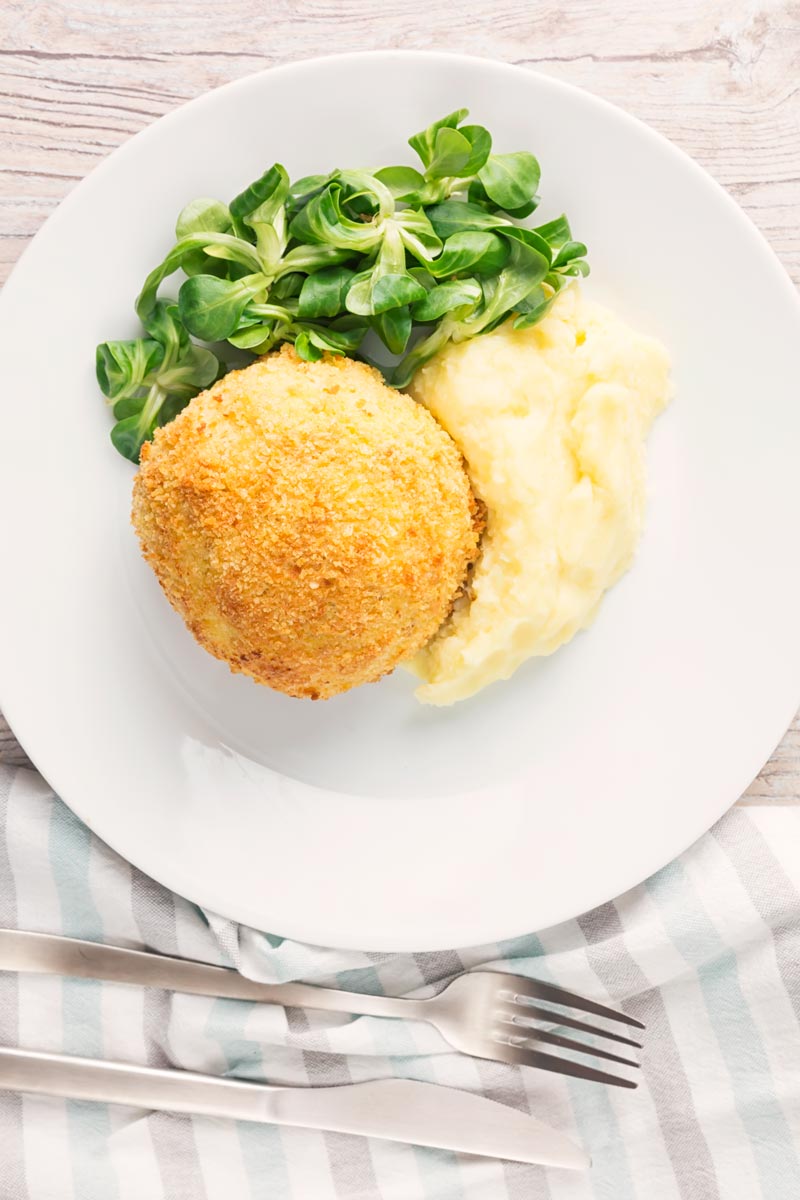Portrait overhead image of a crispy garlic chicken Kiev on a white plate, served with a side salad and mashed potato