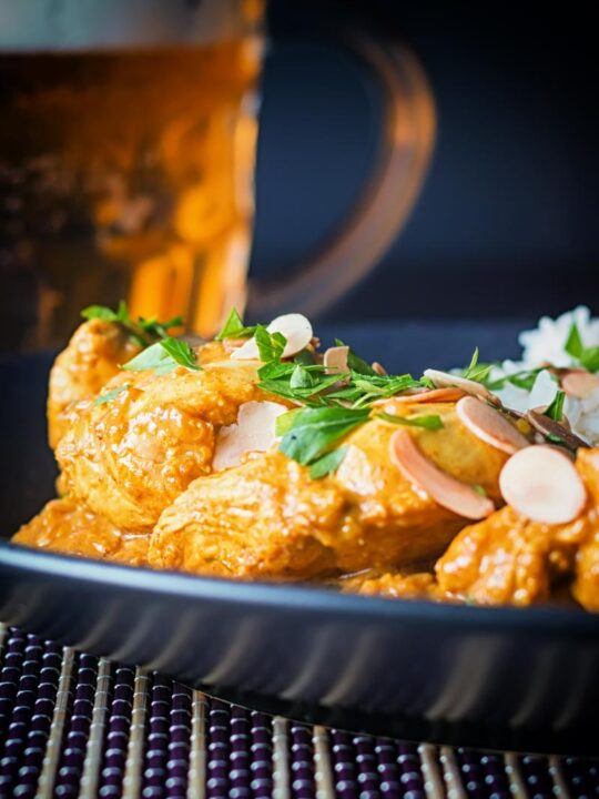 alto close-up imagem de um frango pasanda curry com uma pilha de arroz basmati e amêndoas em flocos em um prato preto com uma cerveja ao fundo