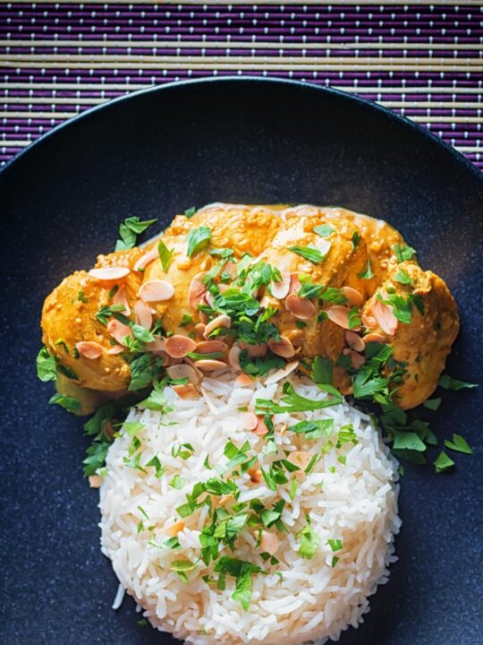 Tall overhead image of a chicken pasanda curry with a pile of basmati rice and flaked almonds on a black plate