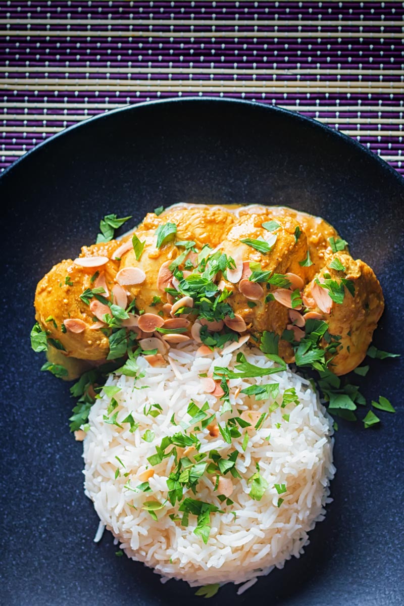 Tall overhead image of a chicken pasanda curry with a pile of basmati rice and flaked almonds on a black plate