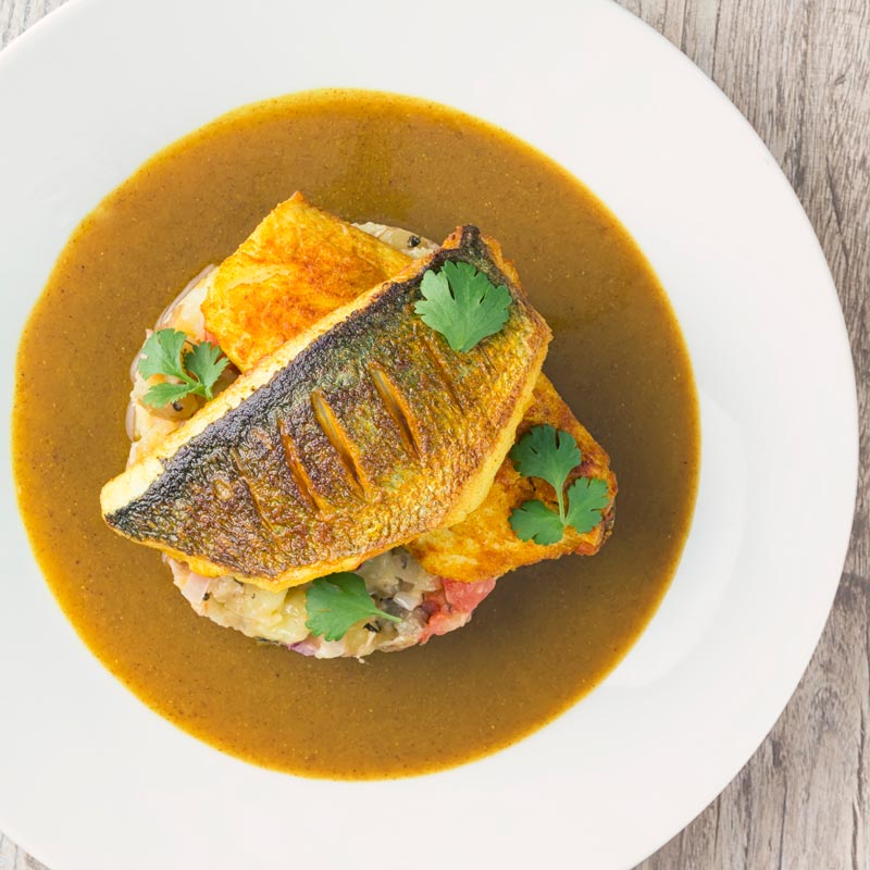 Square overhead image of a deconstructed fish curry with turmeric sea bream fillets on fenugreek potatoes with a curry sauce served on a white plate