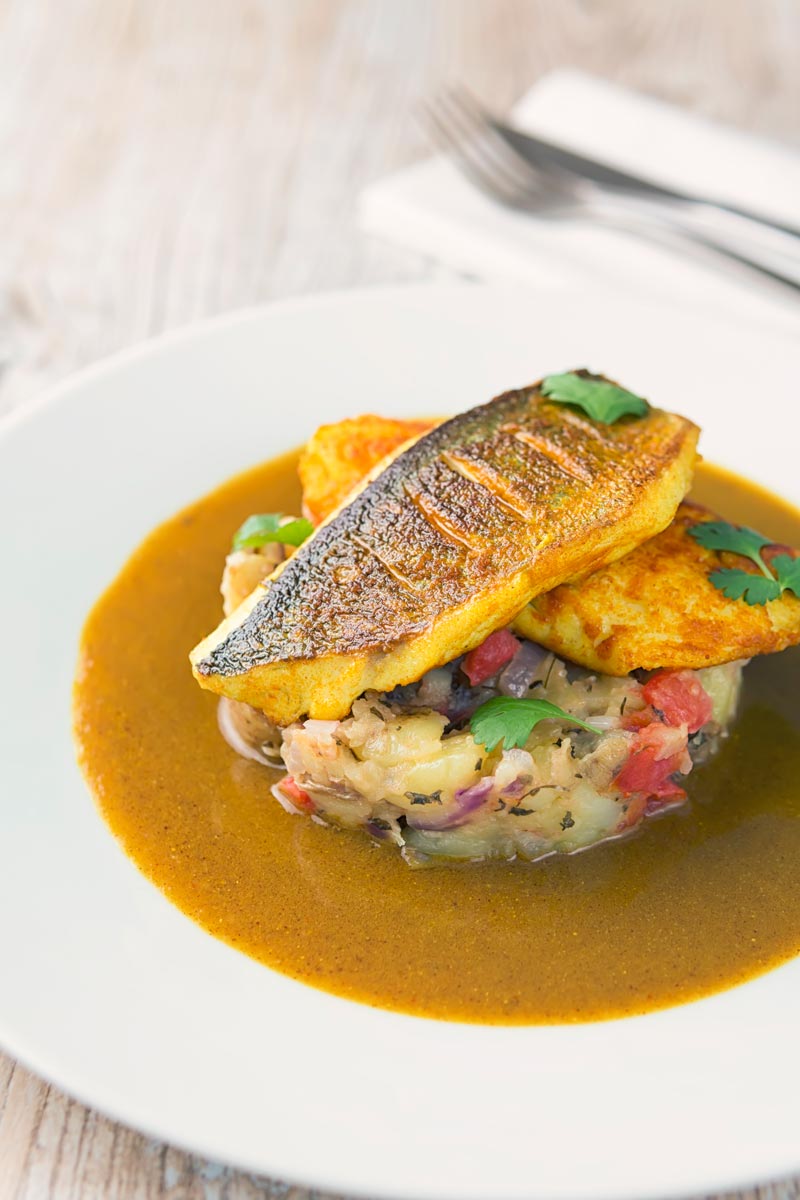 Landscape image of a deconstructed fish curry with turmeric sea bream fillets on fenugreek potatoes with a curry sauce served on a white plate