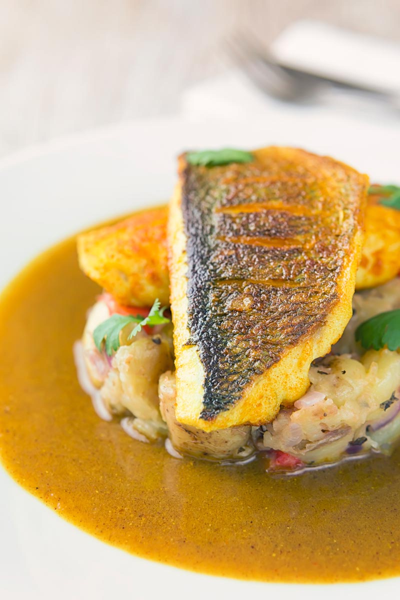 Landscape close up image of a deconstructed fish curry with turmeric sea bream fillets on fenugreek potatoes with a curry sauce served on a white plate