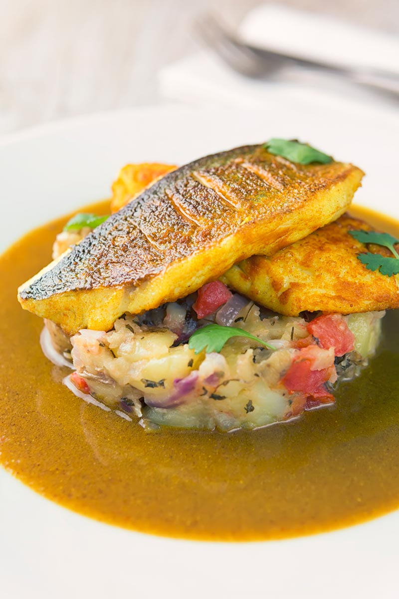 Landscape image of a deconstructed fish curry with turmeric sea bream fillets on fenugreek potatoes with a curry sauce served on a white plate