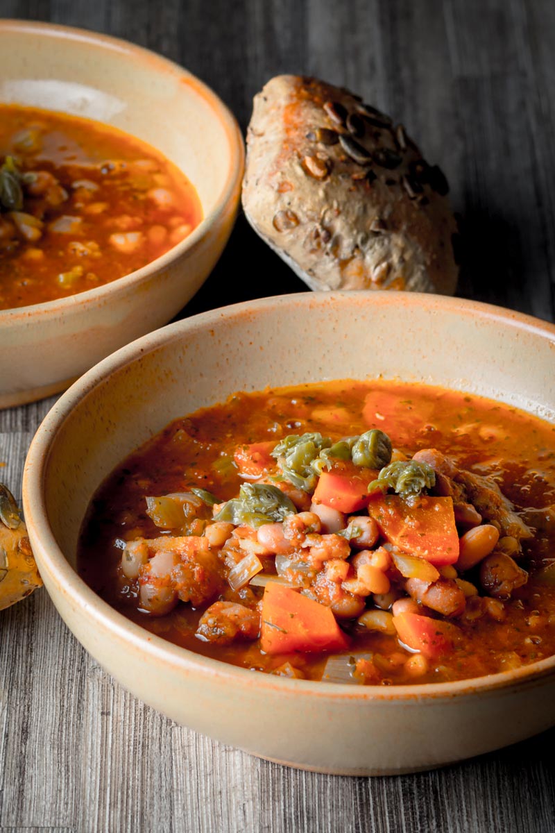Portrait image of two bowls of tomato and bean soup in earthenware bowls served with a seeded bread roll