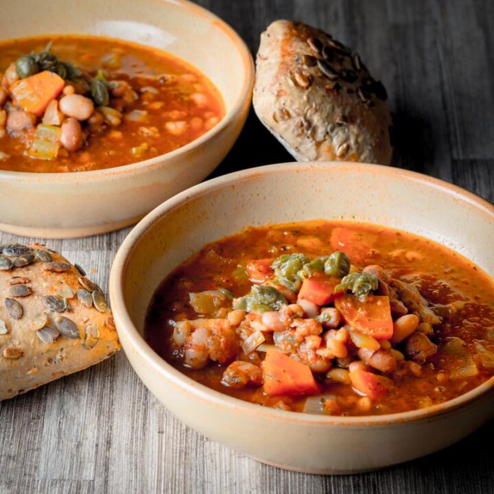 Square image of two bowls of crockpot bean soup in earthenware bowls served with a seeded bread roll