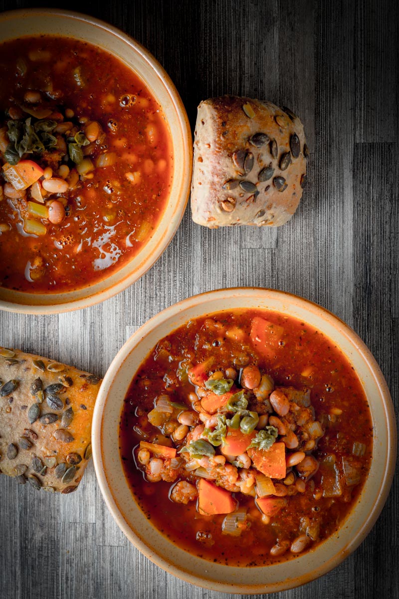 Overhead portrait image of two bowls of tomato and bean soup in earthenware bowls served with a seeded bread roll