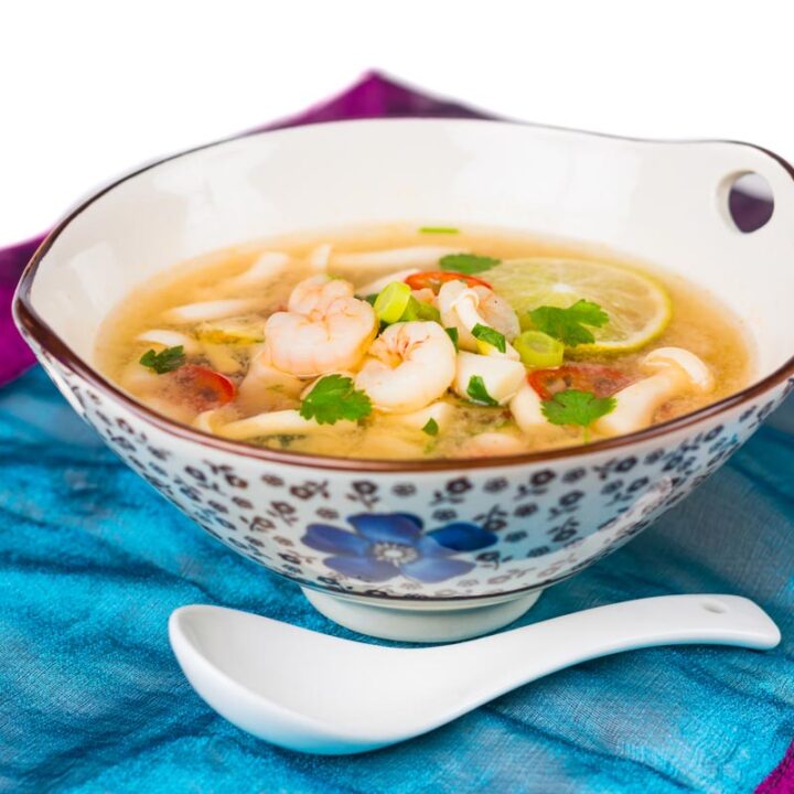 Square image of a hot and sour shrimp soup with mushrooms and chili and lemon in a clear broth in an Asian style bowl decorated with a blue flower