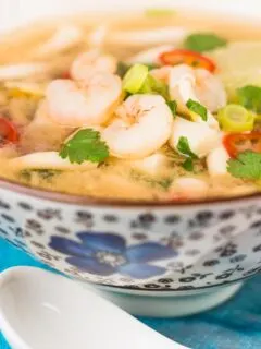 Close up tall image of a hot and sour shrimp soup with mushrooms and chili and lemon in a clear broth in an Asian style bowl decorated with a blue flower
