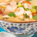 Close up tall image of a hot and sour shrimp soup with mushrooms and chili and lemon in a clear broth in an Asian style bowl decorated with a blue flower with text