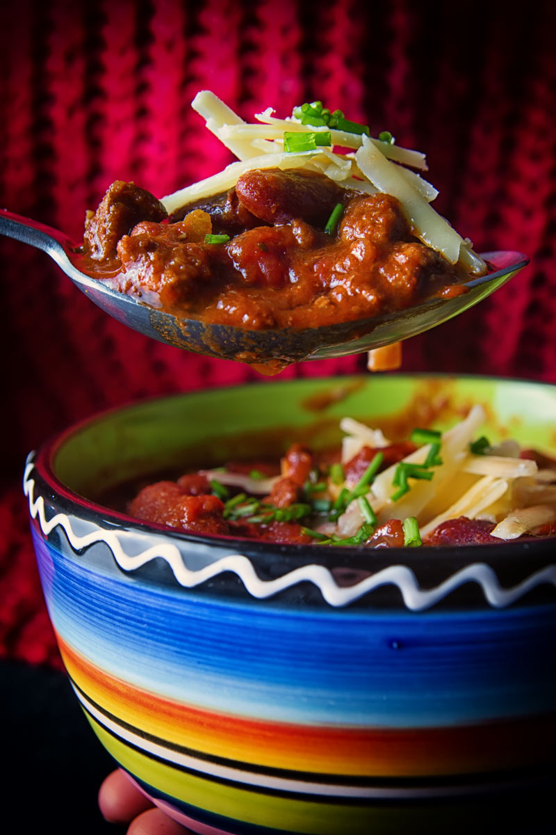 Portrait image of chili con carne topped with sour cream, cheese and chives in a colourful bowl with the dine taking a spoonful