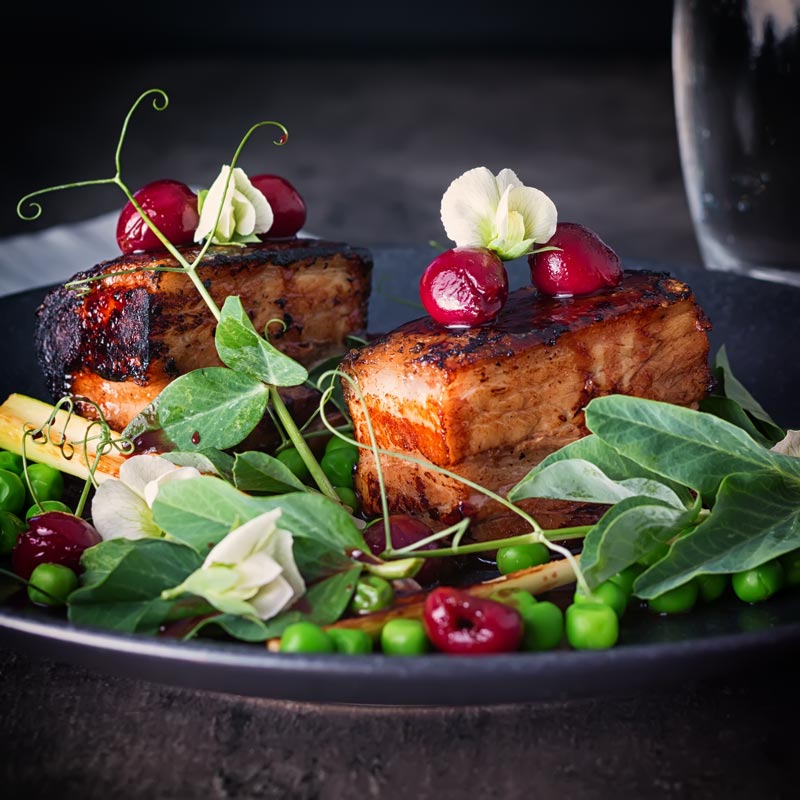 2 slices of pressed pork belly with cherries, peas and pea shoots swerved on a black plate