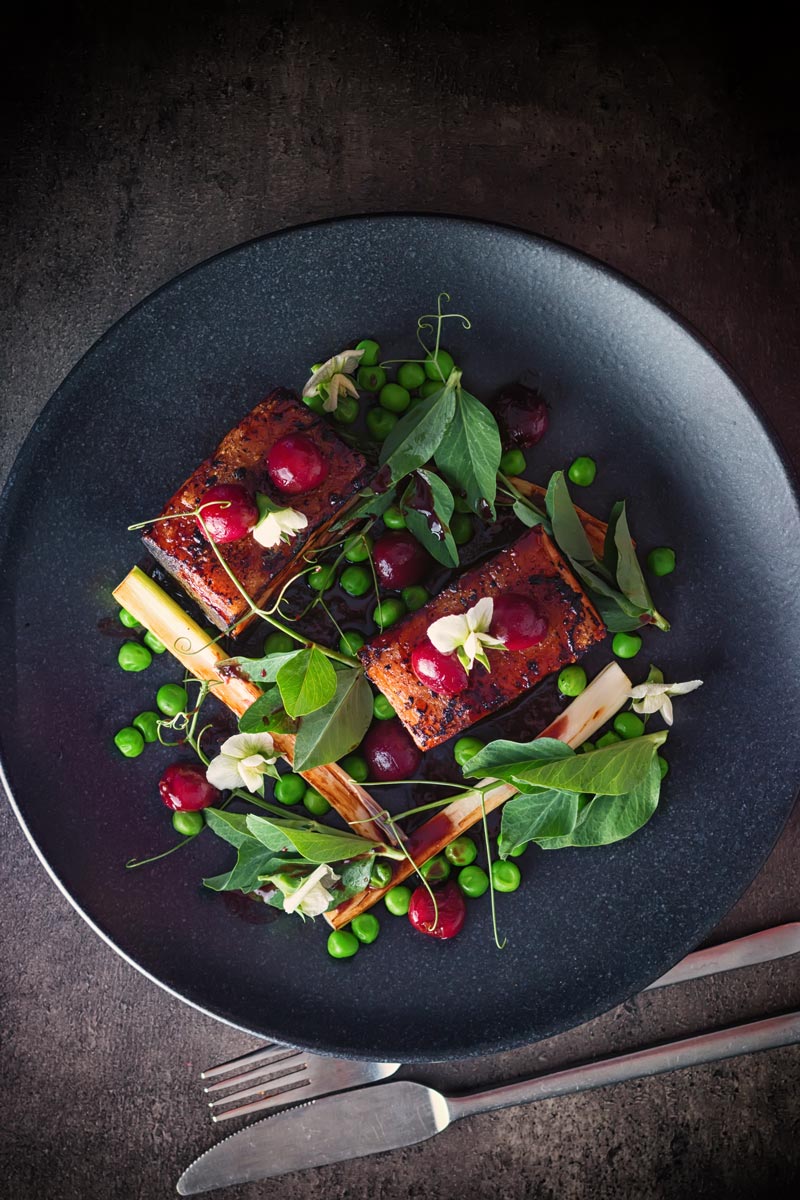 Pressed pork belly with cherries, peas and pea shoots swerved on a black plate photographed from above.