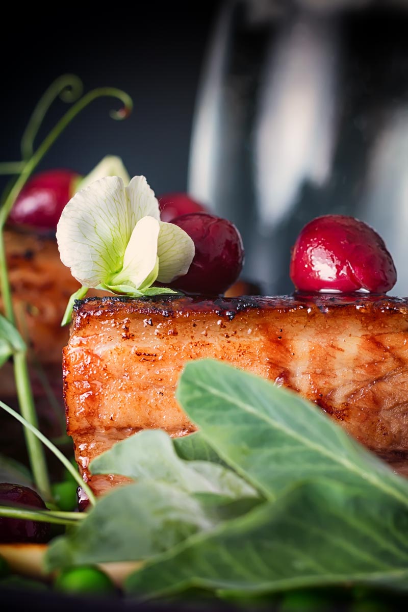 Close up of pressed pork belly with cherries, and pea flowers against a dark backdrop