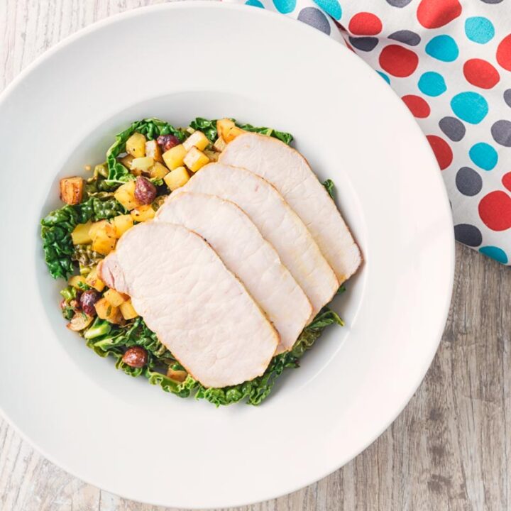 Square image of mustard roast pork on a bed of cabbage and golden fried potatoes and hazelnuts in a white bowl and spotted lined photographed from above