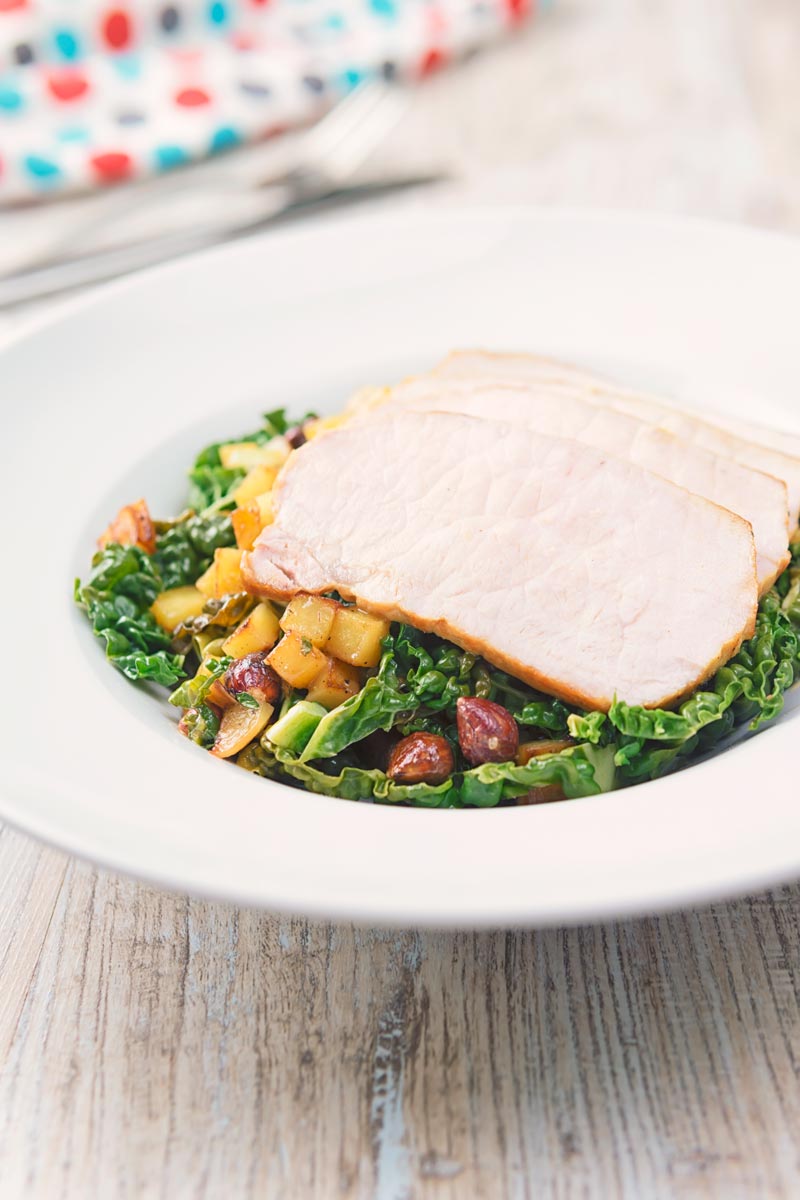 Tall image image of mustard roast pork on a bed of cabbage and golden fried potatoes and hazelnuts in a white bowl and spotted linen