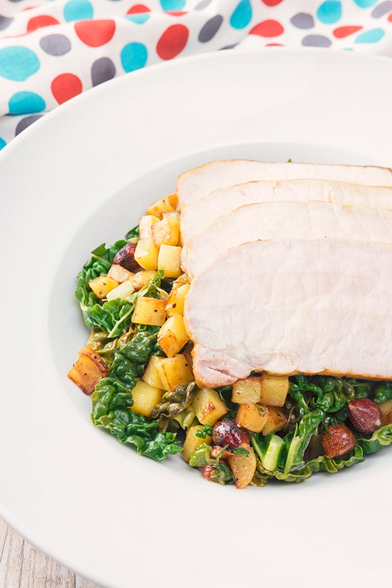 Tall close up image of mustard roast pork on a bed of cabbage and golden fried potatoes and hazelnuts in a white bowl and spotted linen