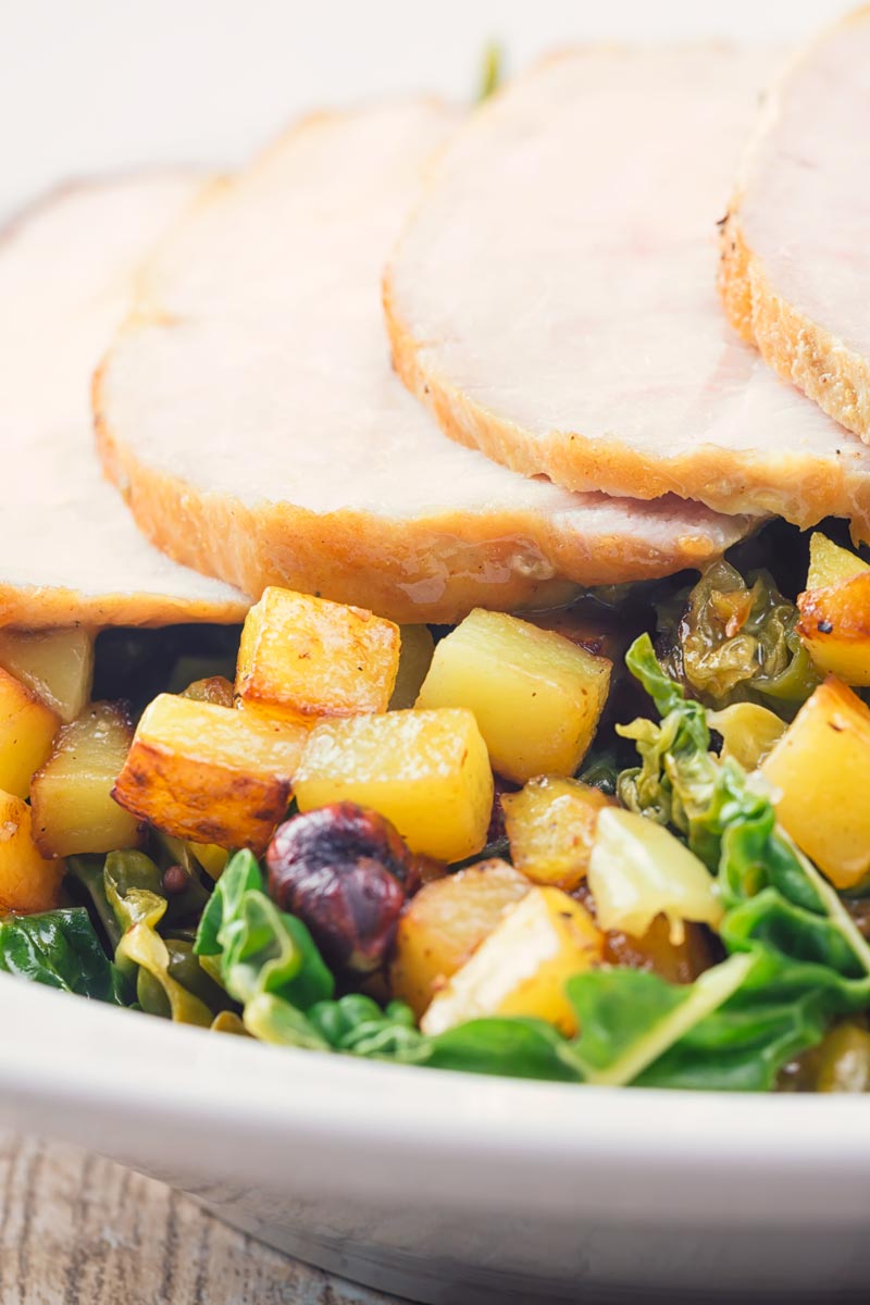 Tall close up image of golden fried potatoes with hazelnuts on a bed of cabbage with roast pork loin in the background