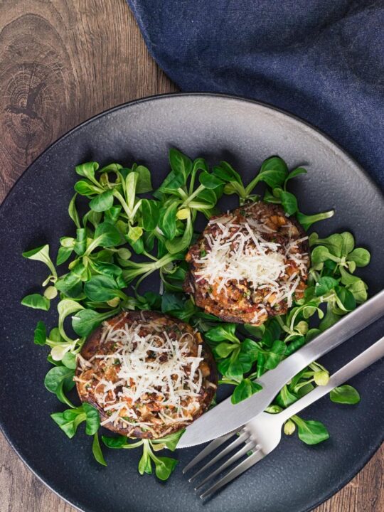 Tall image taken from above of  minced beef stuffed mushrooms topped with Parmesan cheese served on a black plate with salad leaves