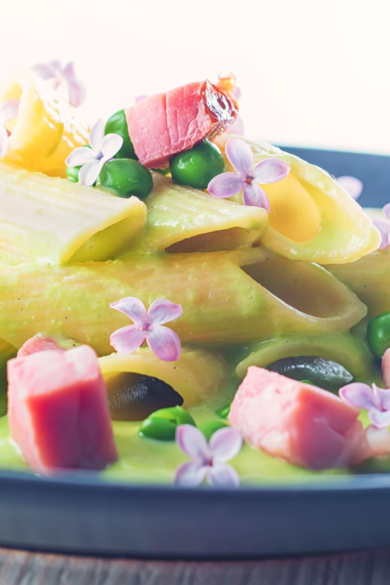 Portrait close up image of pea and Ham pasta with a pea puree, fresh peas, chunks of ham and lilac edible flowers
