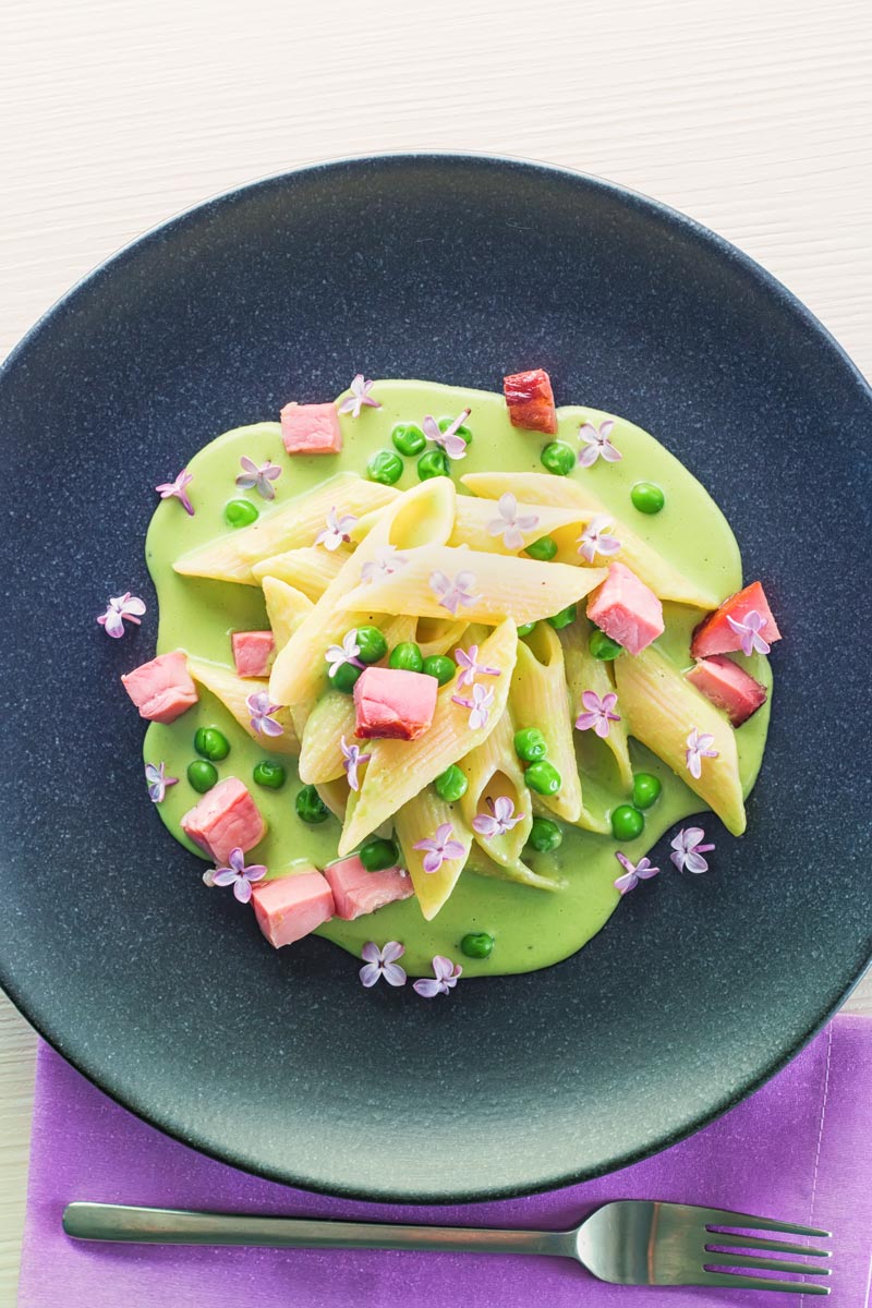 Portrait overhead image of pea and Ham pasta with a pea puree, fresh peas, chunks of ham and lilac edible flowers