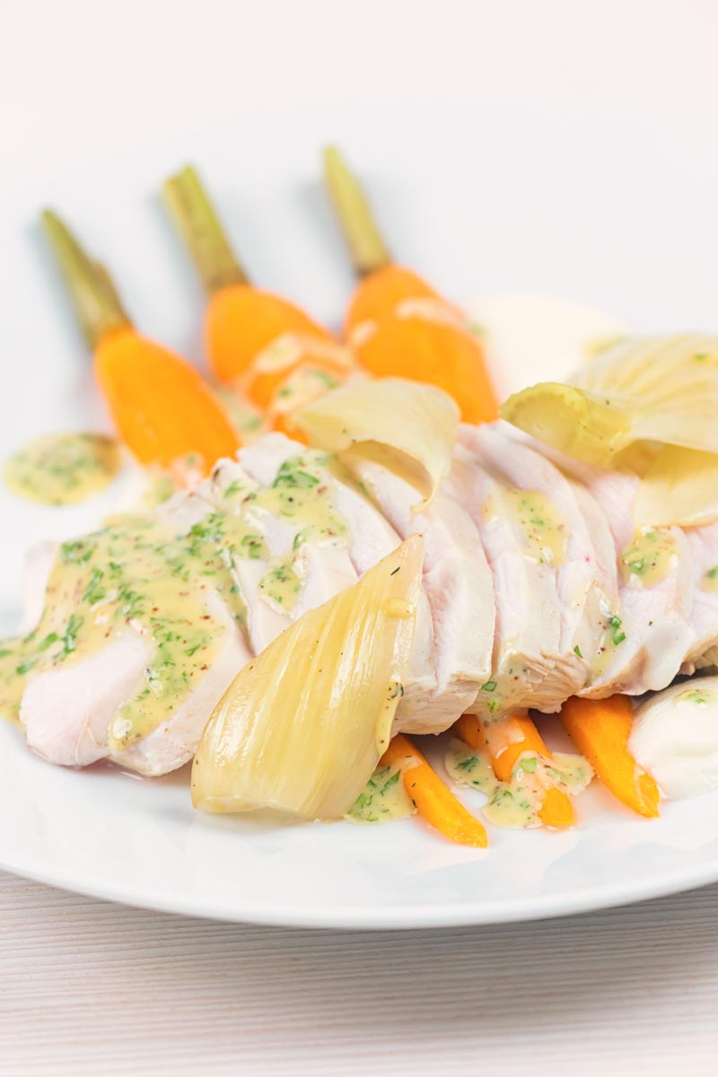 Portrait image of a sliced poached chicken breast on a white plate with carrots, fennel and kohlrabi puree and a mustard sauce