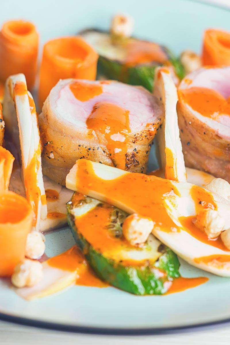 Close up portrait image image of roast pork tenderloin wrapped in bacon served with pickled carrots, seared cucumber and pickled fennel and a harissa based dressing