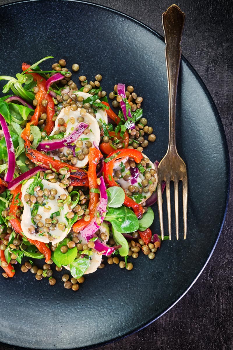 Overhead image of a puy lentil salad with soft goats cheese, red onion and roasted red peppers on a black plate with a vintage fork