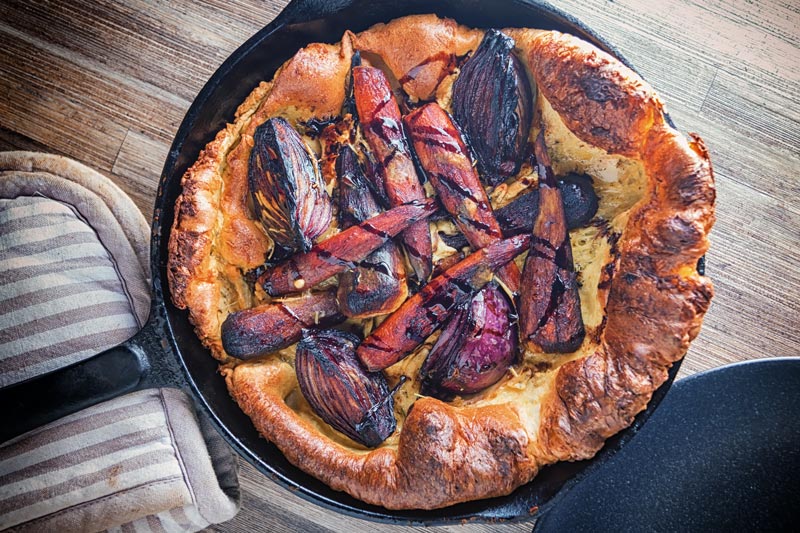 Landscape overhead image of a vegetarian toad in the hole featuring roasted vegetables instead of sausages presented and cooked in a cast iron skillet against a wooden background