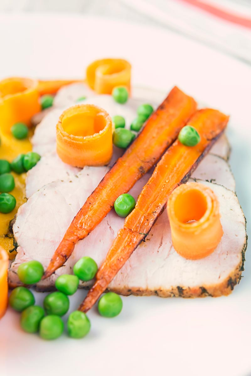 Tall image of roasted pork loin with peas, pickled carrot curls and roasted carrots