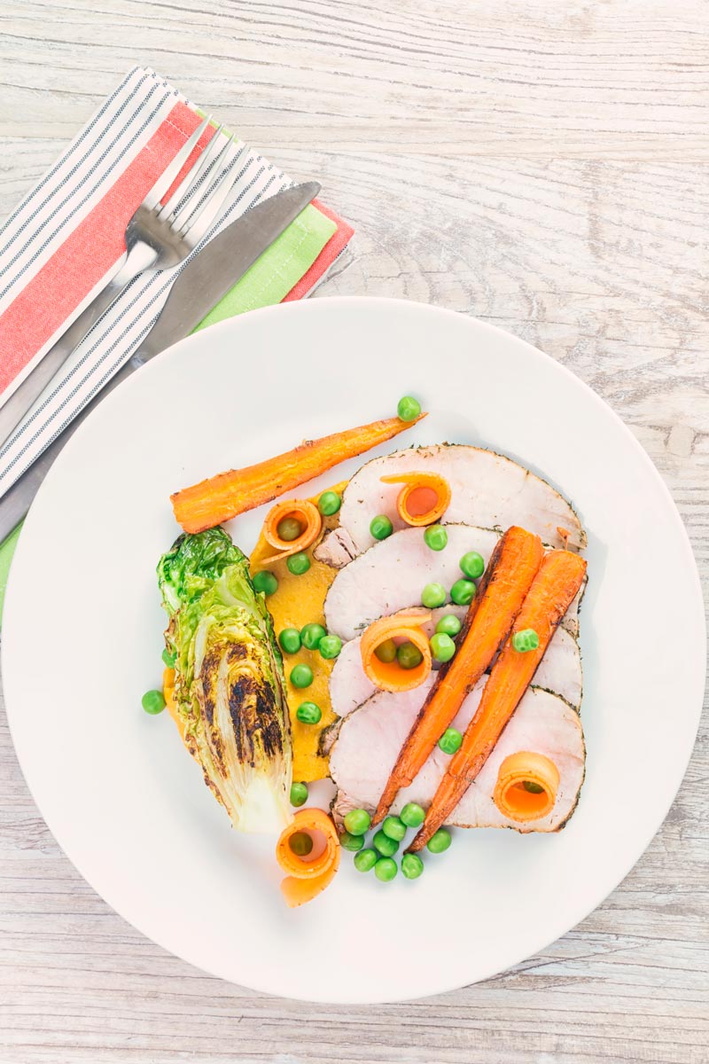 Tall image taken from above of roasted pork loin slices with peas, seared lettuce, pickled carrot curls, carrot puree and roasted carrots