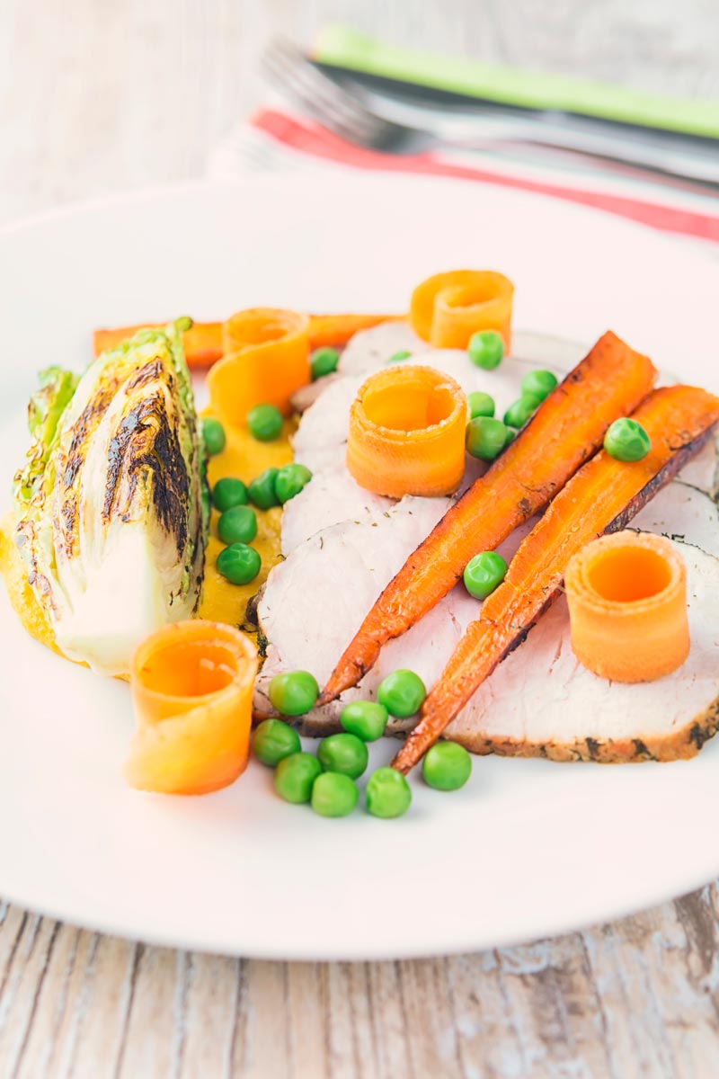 Tall image of roasted pork loin slices with peas, seared lettuce, pickled carrot curls, carrot puree and roasted carrots