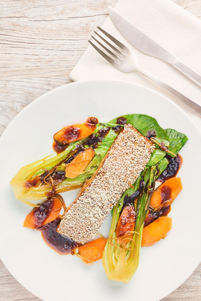 Portrait overhead image of a sesame salmon fillet served with ginger bok choi and carrots on a white plate