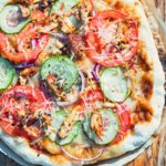 Tall image of a grilled pizza featuring tomato, red onion, cucumber and bbq chicken on a wooden chopping board