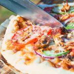 Tall image of a grilled pizza featuring tomato, red onion, cucumber and bbq chicken being cut on a wooden chopping board