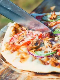 Tall image of a grilled pizza featuring tomato, red onion, cucumber and bbq chicken being cut on a wooden chopping board