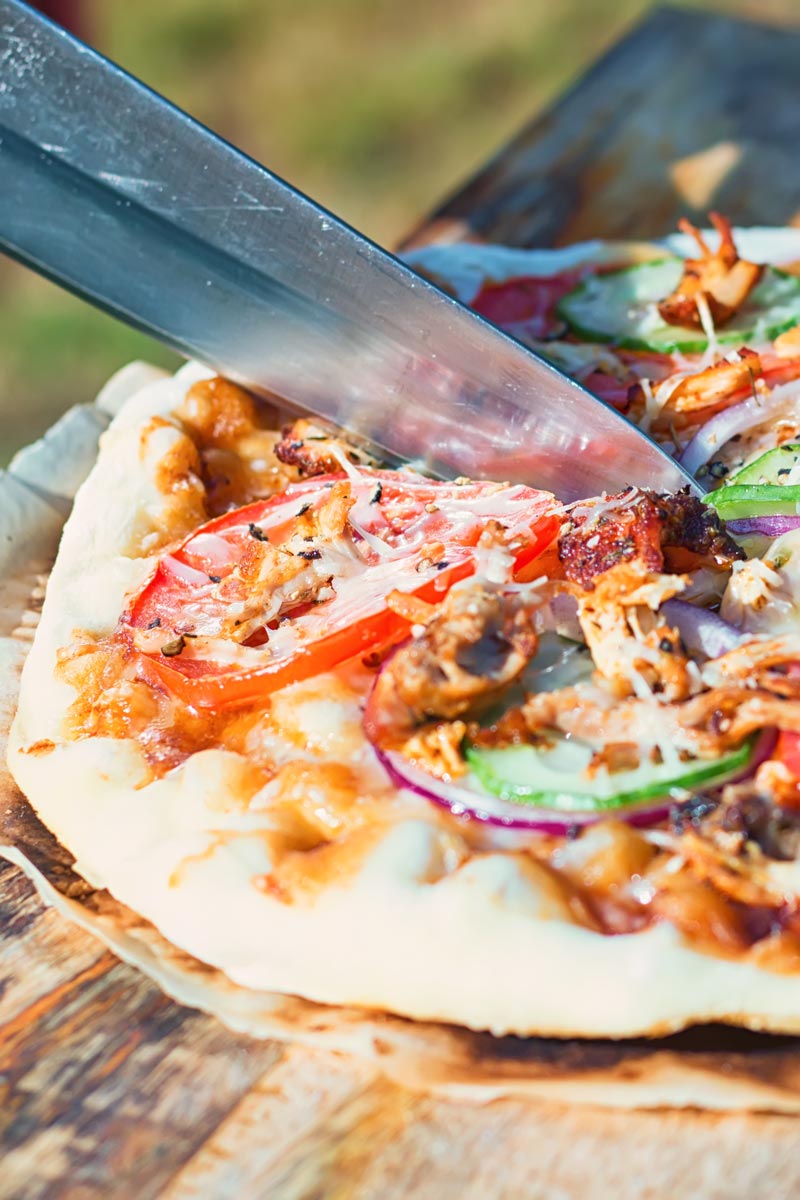 Tall image of a grilled pizza featuring tomato, red onion, cucumber and bbq chicken being cut on a wooden chopping board