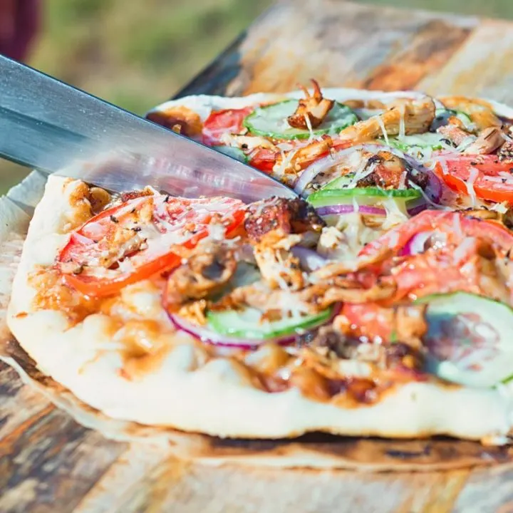 Square image of a grilled pizza featuring tomato, red onion, cucumber and bbq chicken being cut on a wooden chopping board