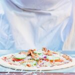 Tall image of a raw pizza being seasoned before being cooked