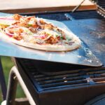 Tall image of a raw pizza being transferred to to a pizza steel on a bbq to become a grilled pizza