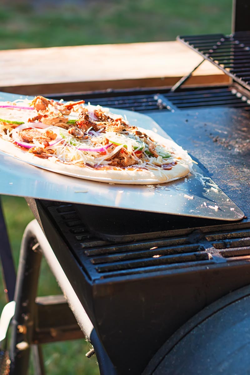 Tall image of a raw pizza being transferred to to a pizza steel on a bbq to become a grilled pizza