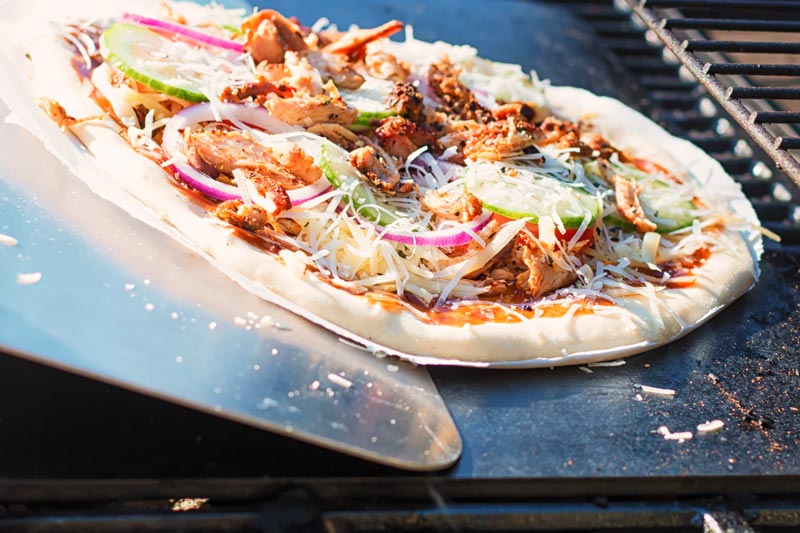 Landscape image of a raw pizza being transferred to to a pizza steel on a bbq to become a grilled pizza