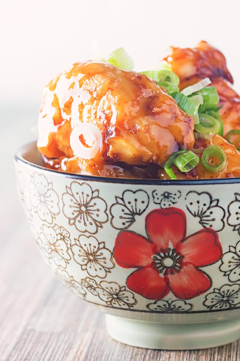 Portrait close up image of Chicken with Honey and Soy with a shiny glaze served in an Asian style bowl decorated with a red flower set against a white backdrop