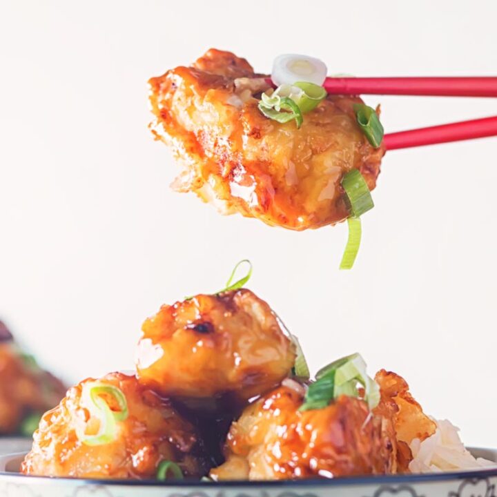 Close up square image of honey and Soy chicken with a shiny glaze being picked up by the diner with red chopsticks set against a white backdrop