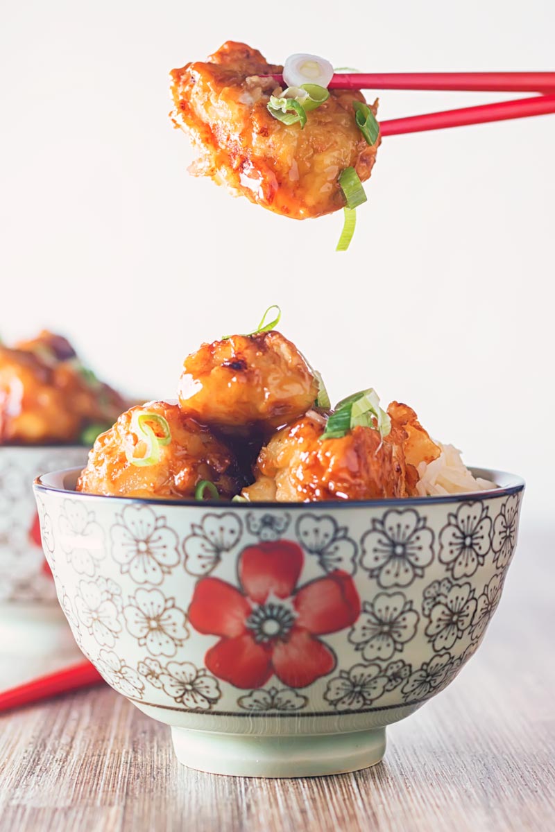 Portrait image of Chicken with Honey and Soy with a shiny glaze being picked up by the diner with red chopsticks set against a white backdrop